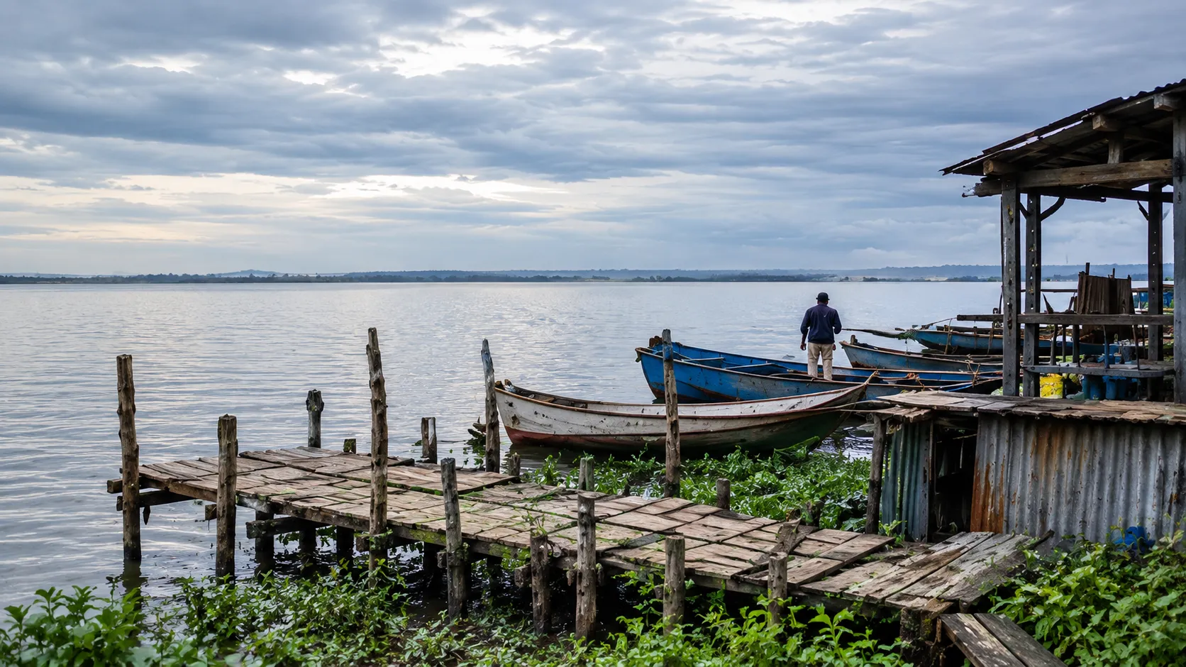 Illustrative visualisation: a working lakeside edge at Kisumu in early morning — a weathered timber jetty, moored fishing boats and corrugated iron structures, with flat grey water and low cloud beyond